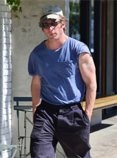 Jeremy Allen White showing off his large arms in a blue t-shirt. He is wearing a baseball cap and glasses